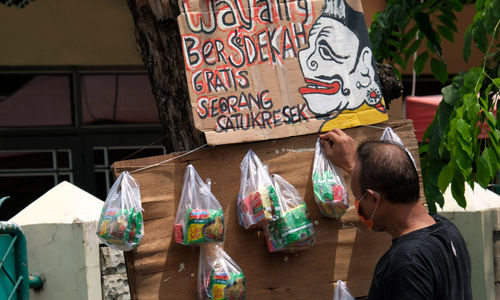 <p>Seniman wayang uwuh Iskandar Hardjodimuljo menggantungkan paket bahan makanan di batang pohon yang akan dibagikan kepada warga yang melintas di Jalan Cawang Baru Utara, Jakarta Timur, Kamis (16/4/2020). Aksi bertajuk “Wayang Bersedekah” ini merupakan inisiatif Iskandar dalam menanggapi kondisi pandemi corona. Bahan makanan yang dibagikan kepada warga di dapat dari hasil penjualan wayang berbahan sampah daur ulang. Dalam sehari rata-rata sebanyak 20 paket makanan dibagikan untuk warga yang kesulitan akibat dampak pandemi COVID-19 saat ini. Foto: Ismail Pohan/TrenAsia</p>
