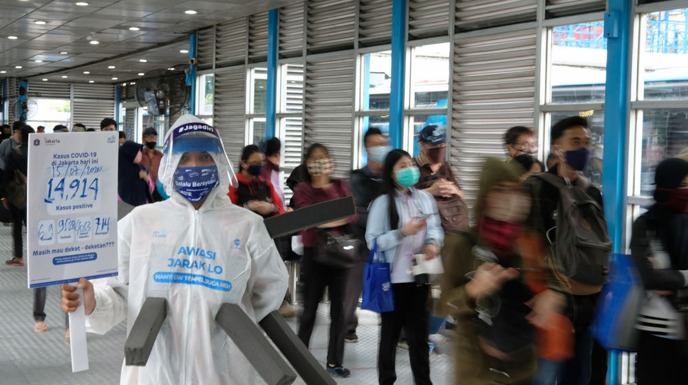 <p>Maskot ‘Manusia Covid’ dari anggota komunitas pecinta Transjakarta mengenakan hazmat dengan membawa poster berisi informasi jumlah kasus positif di Jakarta saat melakukan sosialisasi protokol kesehatan di Halte Transjakarta Harmoni, Jakarta, Rabu, 15 Juli 2020. Sosialisasi tersebut untuk mengingatkan penumpang Transjakarta agar selalu menaati protokol kesehatan saat menggunakan transportasi publik untuk memutus rantai penyebaran COVID-19. Foto: Ismail Pohan/TrenAsia</p>