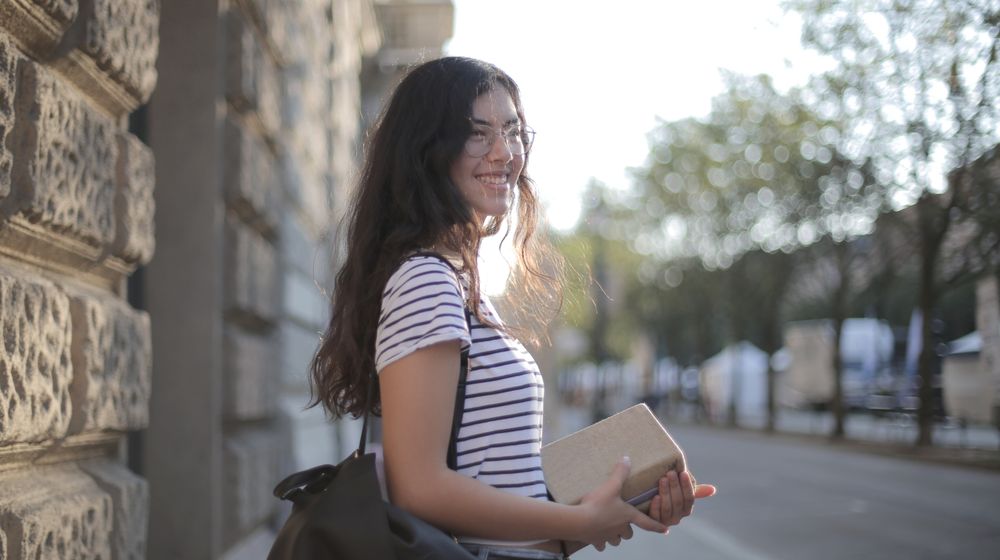 Photo by Andrea Piacquadio: https://www.pexels.com/photo/optimistic-ethnic-woman-with-stack-of-books-in-city-3808181/