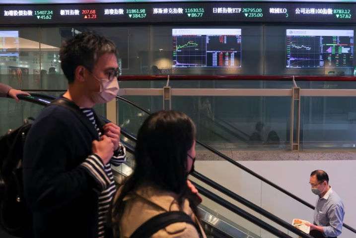 Orang-Orang yang Memakai Masker Wajah Lewat di Depan Layar yang Menampilkan Data Perdagangan (Reuters/Annabelle Chih)