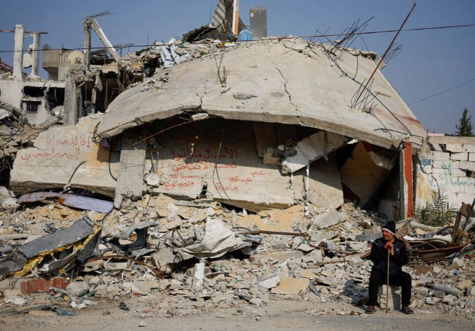 Seseorang duduk di samping tulisan yang dilukis di dinding di tengah puing-puing rumah keluarga, yang hancur dalam serangan mematikan Israel di tengah konflik yang sedang berlangsung antara Israel dan Hamas (Reuters/Mohammed Salem)