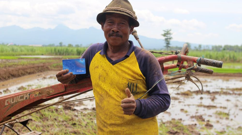 Petani memegang buku tabungan BRI di sawah sambil mengacungkan jempol setelah menerima bantuan BLTS Kesra.