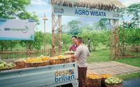 Dua warga sedang bertransaksi buah-buahan di area Agro Wisata Desa Brilian dengan papan program BRI Menanam terlihat di latar belakang.