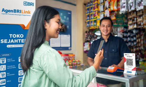 Perempuan melakukan transaksi digital di warung yang menjadi AgenBRILink, sementara petugas warung berdiri di belakang meja dengan alat pembayaran dan papan layanan BRI di sekitarnya.