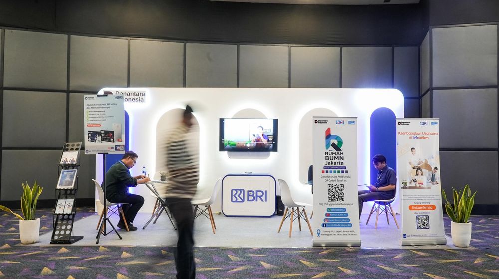 Booth BRI dengan banner program Rumah BUMN dan LinkUMKM, dua orang duduk berkonsultasi, serta seorang pengunjung berjalan melewati area pameran dalam sebuah acara publik.
