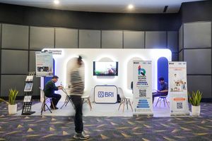 Booth BRI dengan banner program Rumah BUMN dan LinkUMKM, dua orang duduk berkonsultasi, serta seorang pengunjung berjalan melewati area pameran dalam sebuah acara publik.