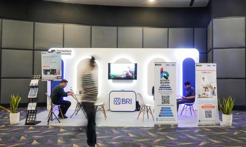 Booth BRI dengan banner program Rumah BUMN dan LinkUMKM, dua orang duduk berkonsultasi, serta seorang pengunjung berjalan melewati area pameran dalam sebuah acara publik.