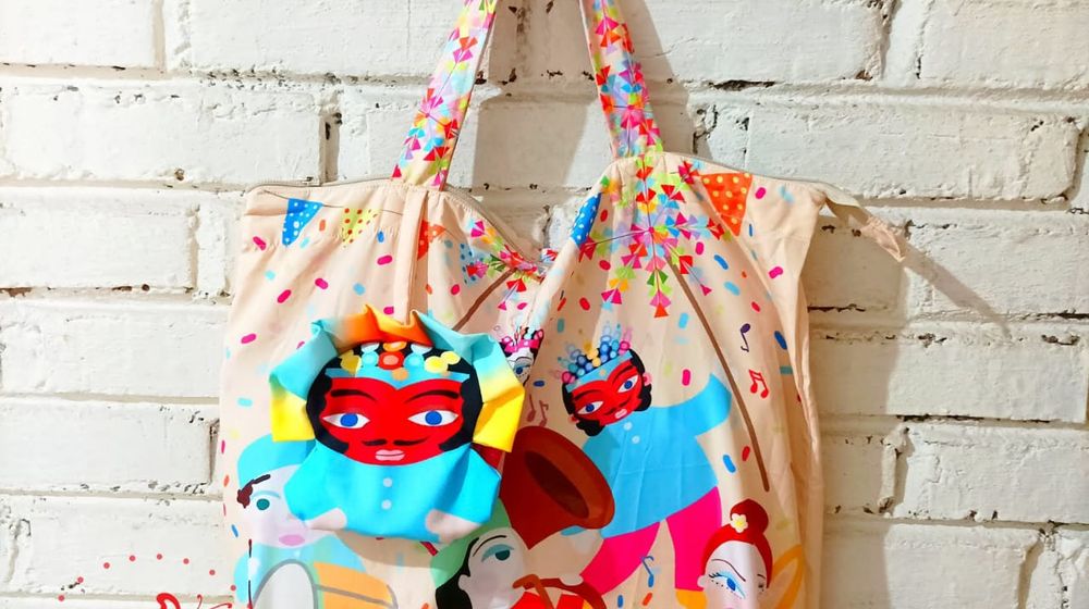 Tote bag bermotif ilustrasi budaya Betawi dengan karakter ondel-ondel dan musisi tradisional, digantung di dinding bata putih sebagai produk kerajinan Pundi Craft.