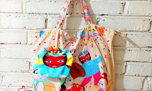 Tote bag bermotif ilustrasi budaya Betawi dengan karakter ondel-ondel dan musisi tradisional, digantung di dinding bata putih sebagai produk kerajinan Pundi Craft.